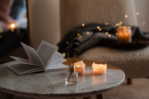 Home liquid fragrance with scented candle and open paper book over christmas glow lights and chair in bedroom. Cozy hygge aroma atmosphere.