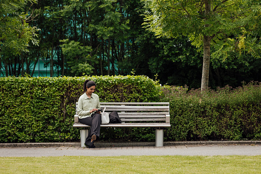 A full shot of a businesswoman typing on her mobile phone looking down at it whilst being sat down on a bench. She is dressed smartly in a shirt, sitting in a business park. She is based in the North East of England