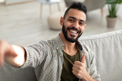 Happy young Arab guy taking selfie and showing thumb up gesture, smiling at mobile phone camera, sitting on sofa at home. Portrait of positive middle Eastern man making photo of himself indoors