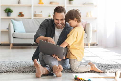 Modern kids interests. Cute little boy watching his daddy working on laptop, cheerful man showing son how to work online, sitting on floor at home