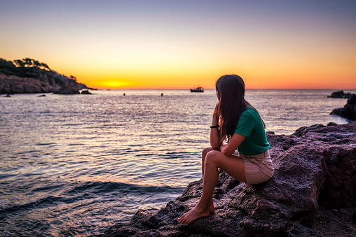 Happy young female contemplating the scenic sunset seated on a rock at costa brava
