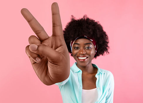 Happy young black woman showing big victory v sign on pink studio background. Cheery African American lady making peace gesture, smiling at camera. Selective focus