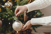 Hands decorating christmas tree with stylish baubles in atmospheric festive room. Merry Christmas! Winter holidays preparation. Woman in cozy sweater putting vintage baubles on tree