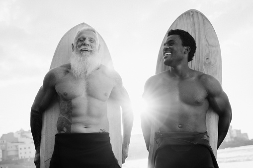 Multiracial senior and young men surfers having fun after surf training at the beach - Travel vacation - Soft focus old man face - Black and white editing.