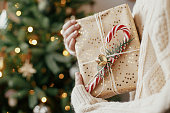 Merry Christmas! Stylish christmas gift in hands against christmas tree with lights.Woman in cozy sweater holding wrapped present with candy cane in festive room. Holiday banner, space for text