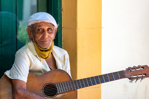 Havana, Cuba - August 26, 2022: Portrait of a street musician in Old Havana district. He slightly smiles and holds an old acoustic guitar.