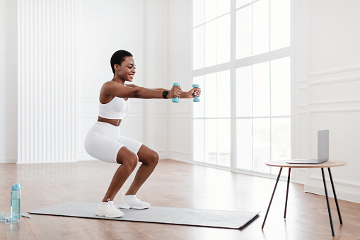 Positive black lady exercising and squatting with two dumbbells in front of pc on tea table at home, smiling fitness blogger recording video for her followers having personal virtual training