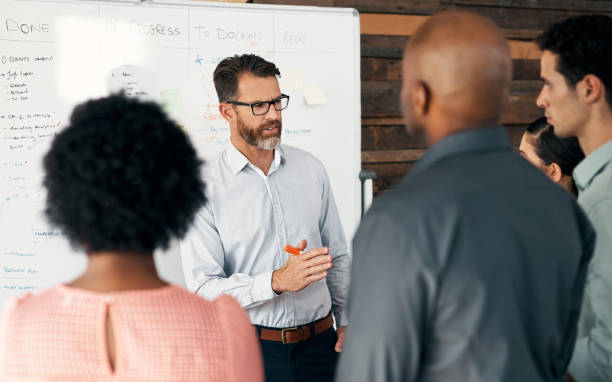 istock Businessman, whiteboard and office mentor training diversity workers, employees and designers in project management with schedule. Talking manager, ceo or leadership in teamwork coaching presentation 1434120796