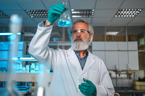 Waist up portrait of a mature male pharmaceutical researcher wearing  protective gloves and glasses, lab equipment in blurred foreground. Holding up a condenser with blue liquid and examining it, small glass jar in other hand. Looking away.