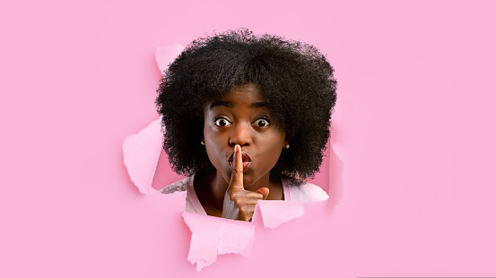Shocked millennial black female looks into hole in pink paper, shows finger near lips, sign of silence. Amazing interesting offer and advertising, sales and facial expressions, gossip and secret