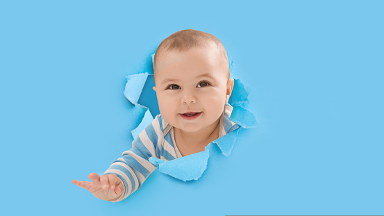 Smiling cute adorable european little kid climbs through hole on blue paper, copy space, panorama. Happy childhood, healthy child, family and people. Portrait of newborn baby interested something