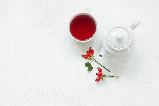 Rosehip glass cup of herbal tea medicinal plants with rosehip branch and ceramic teapot on white table. Dogrose winter tea background