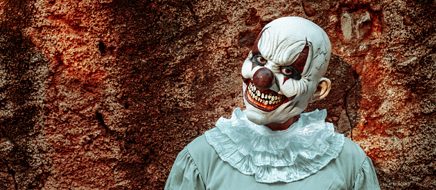 an evil clown, wearing a gray costume with a white ruff, stares at the observer with a scary smile, in front of the rusty wall of an abandoned house, in a panoramic format to use as web banner