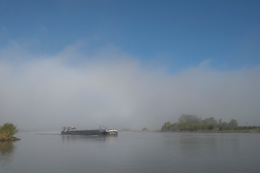 Freight ship sailing on the river IJssel during a misty morning sunrise in the IJsseldelta landscape along the river IJssel during a autumn morning in Overijssel, The Netherlands.