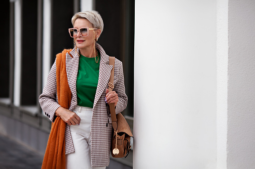 Stylish woman short hairdo wearing double-breasted jacket, warm scarf, bag, glasses with chain looks away, waiting for somebody. Autumn or spring fashion personal style