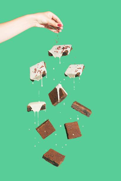 istock Minimal composition with chocolate brownies covered
with white cream floating against green background. Female hand pouring crumbs on cookies. Tasty dessert concept. Abstract food visual. 1434133363