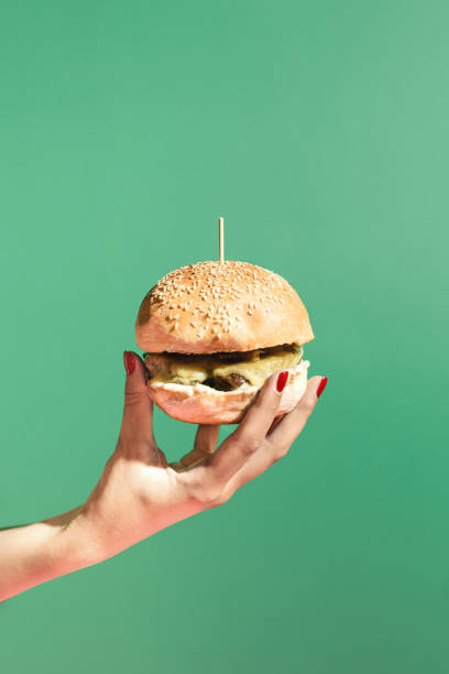 istock Modern scene with womans hand holding a burger against green background. Minimal composition with copy space. Fast food and street food concept. 1434133443