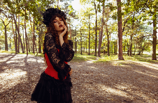 A young brunette woman in a carnival costume with a wreath on her head with her eyes closed stands in a pine forest. Halloween woman portrait in nature. fantasy woman