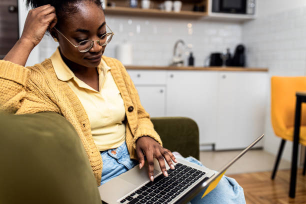 istock Young African American woman sitting on sofa at home studying using laptop. 1434138560