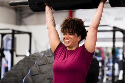 A woman weightlifting a strongman log in a gym in Newcastle upon Tyne, England. She is lifting the log above her head and smiling.