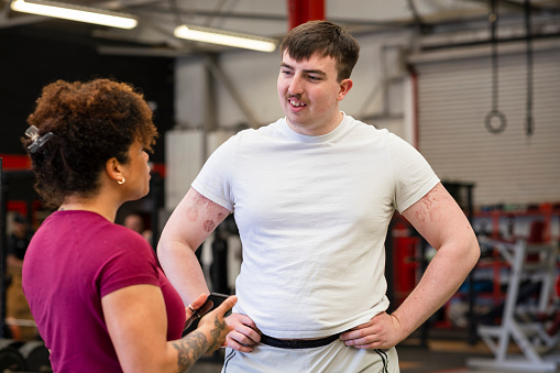 Two gym goers, standing in a gym surrounded by gym equipment in Newcastle upon Tyne, England. They are discussing training exercises with each other and getting ready to train together.