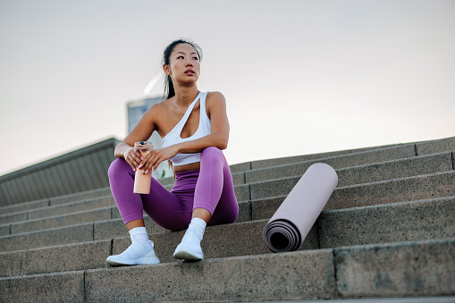 A young Asian female urban athlete is sitting on the steps in the city holding a bottle of water in her hands resting after a workout.