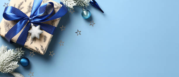 Gift box with blue ribbon bow on a pastel blue background with fir branches and baubles. Christmas banner design, New Year header template.