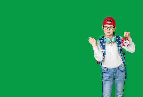 Little happy girl in eyeglasses and headphones with backpack holding alarm clock. A schoolgirl holds alarm clock on green background. Time to go to school. Back to school concept