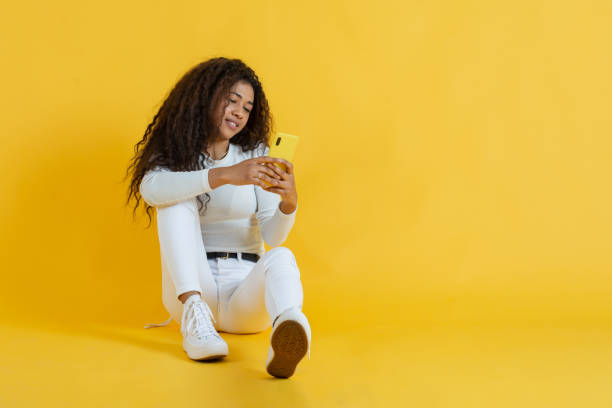 Relaxed young woman sitting on floor using her cell phone on yellow background stock photo