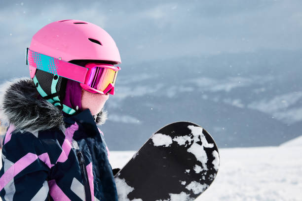snowboarder de niña con casco de equipo y gafas de ropa exterior sosteniendo una tabla de snowboard descansando en la cima de la pista de esquí a la luz del sol. niña de 6-7 años en la estación de esquí en un soleado día de invierno. espacio libre p - 6 7 years lifestyles nature horizontal fotografías e imágenes de stock
