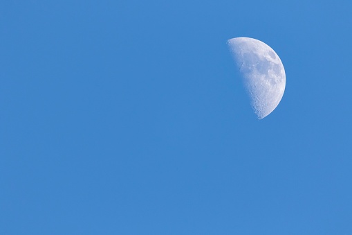 A portrait of a half moon during day time in a blue sky. The craters on the visible have of the satelite planet are well visible.