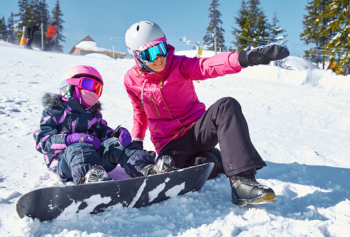 Mother teaches his daughter to snowboard. Woman sits and showing way to ride on ski slope at sunny winter day, travel vacation, landscape mountains background.