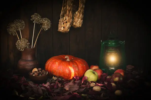 Photo of Autumn authentic rustic set on wooden backdrop with pumpkin, paraffin lamp, nuts, apples, clay jug and bast shoes, low key, selective focus.