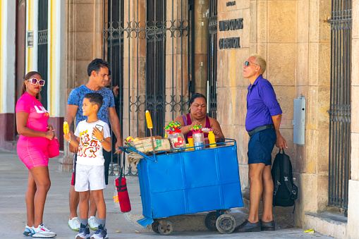 Havana, Cuba - August 15, 2022: Woman selling sweets from a cart on a street corner. A family of a man, woman, and boy is buying sweets from the vendor, and another man with his hands behind his back is standing beside the cart.