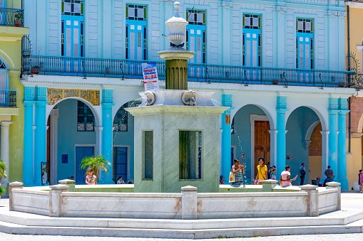 Havana, Cuba - September 16, 2022: People standing and sitting in front of a blue-story building. A fenced monument with a fountain is in front of the building.