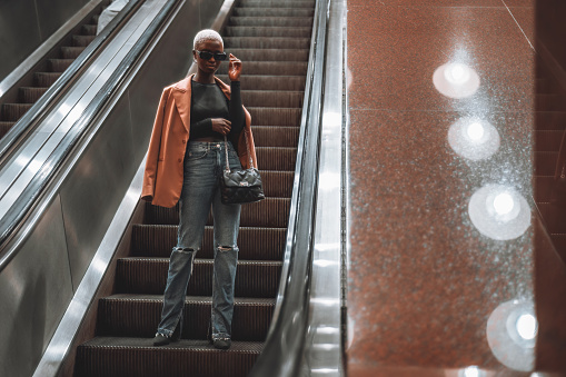 A young fashionable black female on the moving staircase descending to the subway station; a youthful dapper African lady in an overcoat, sunglasses, and painted white hair going down on an escalator