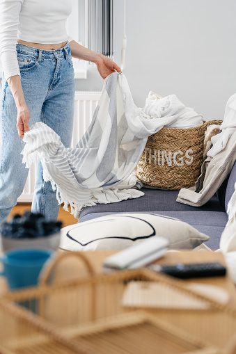 cropped shot of female in jeans take clean bed clothes or linen sheets from wicker basket in living room at apartment, housework concept