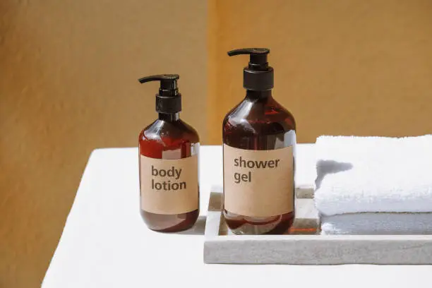 shower gel and body lotion in brown plastic bottles with dispenser, white clean bath towel folded on marble tray at home bathroom on yellow background, personal care concept