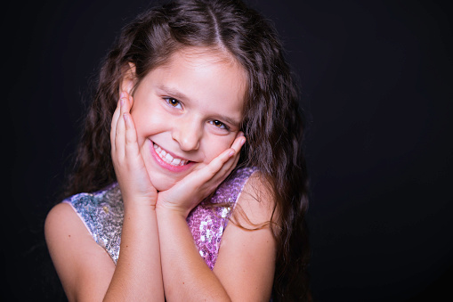 Cute little girl with blonde hair and blue eyes, dressed with a blue navy dress, holds her hands together, smiling she's looking at the camera, the background is blacked, two speedlights were used for the shot.