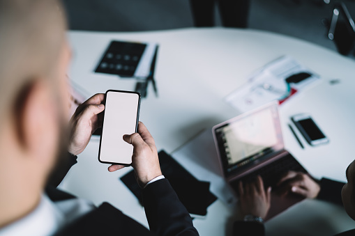 From above anonymous male entrepreneur in formal wear using smartphone with empty screen while standing near table during business conference in office