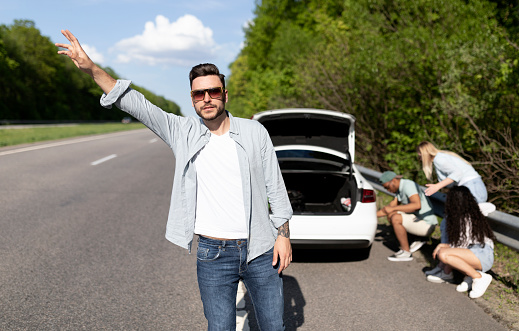 Young Caucasian man trying to wave down car, asking for help, his friends trying to fix broken vehicle, having flat tire on road. Millennial guy hitchhiking on highway, needing emergency assistance