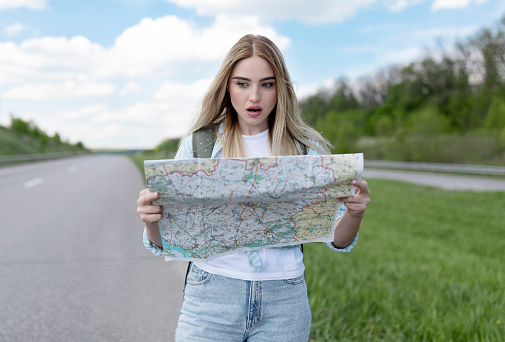 Shocked young woman hitchhiking on road, looking at map in terror, feeling lost while traveling alone by autostop. Millennial blonde lady standing on highway, cannot find her destination
