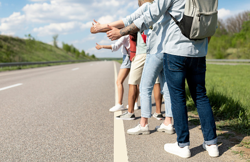 Group of unrecognizable young friends standing along road, making thumb up gesture, hitchhiking for car during road trip, traveling together in summer, closeup. Outdoor vacation, tourism concept