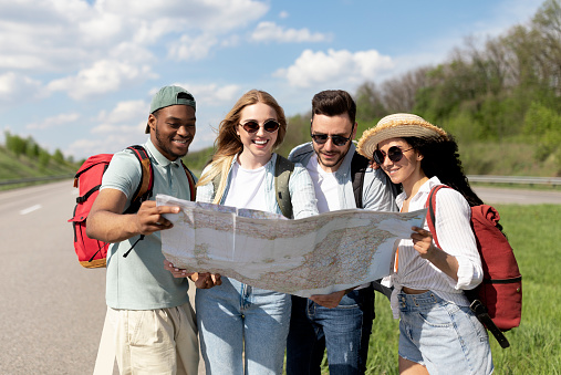 Group of multinational young friends looking at map, choosing route during road trip, hitchhiking on highway. Autostop journey, summer holidays, freedom and friendship concept