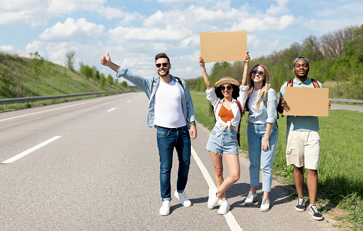 Happy diverse friends standing on highway with empty signs, stopping car, going on trip together, outdoors. Group of multinational hitchhikers traveling by autostop, having fun during road trip
