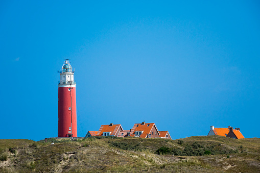 The lighthouse of the island of Texel in the Netherlands against a clear blue sky. The roofs of the surrounding houses peek over the dunes in the foreground.
