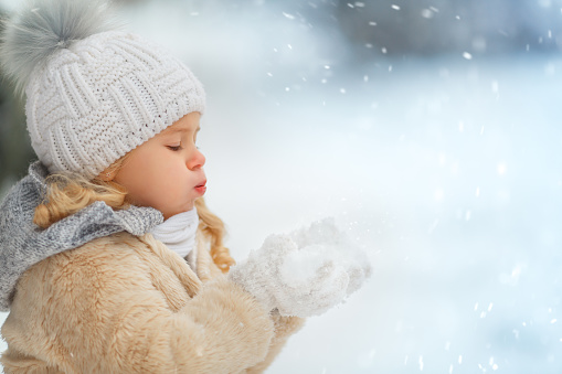 A girl in a coat and hat in winter blowing snow off her hands in a snowy forest. Funny child portrait.