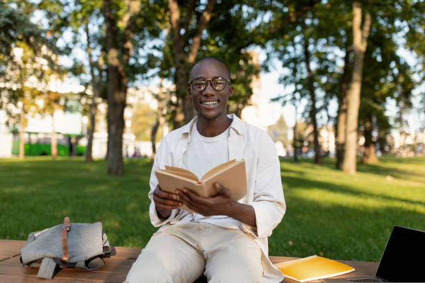 istock Happy black student guy reading interesting book, having rest after classes in park, sitting on bench on sunny day 1434198547