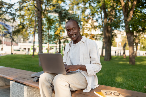 Rest at campus. Happy african american male college student relaxing outdoors with laptop, sitting on bench in park and smiling at camera, copy space