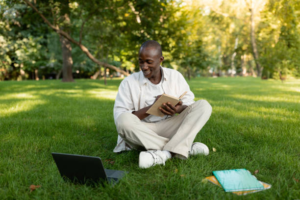 istock Exam study concept. Black male student guy taking notes while watching online lesson on laptop, sitting on lawn in park 1434201104